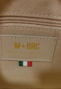 Tessuto beige con un'etichetta in pelle che riporta "M★BRC GENUINE LEATHER MADE IN ITALY" in oro. Presenta un piccolo emblema della bandiera italiana sottostante.