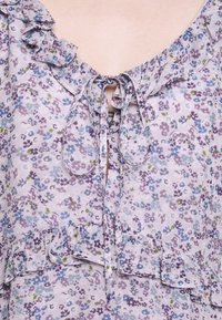 Blouse florale lavande avec petites fleurs bleues et violettes, encolure et ourlet volantés, nouée avec un nœud à l'ouverture devant.