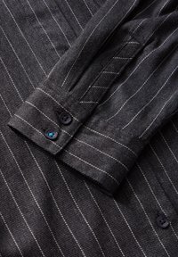 Tissu gris foncé à fines rayures avec gros plan sur un poignet de chemise boutonné comportant deux boutons noirs et de fines rayures verticales blanches.