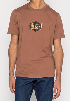 Mężczyzna w brązowej koszulce z krótkim rękawem z wielokolorowym logo Diesel umieszczonym na środku klatki piersiowej, w połączeniu z ciemnoniebieskimi dżinsami.
