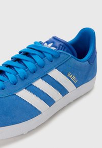 Baskets en daim bleu avec trois rayures blanches et un logo "Gazelle" jaune. Elles présentent un col rembourré et une semelle en caoutchouc texturée.