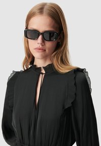 Abito nero con maniche arricciate e scollatura profonda. Modella che indossa occhiali da sole neri a forma rettangolare. Tessuto liscio, design elegante, hardware minimal.