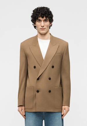 Άνδρας με σγουρά μαλλιά, φορώντας ένα διπλό breasted μπεζ σακάκι πάνω από λευκό πουκάμισο και μπλε τζιν, στέκεται μπροστά σε απλό φόντο.