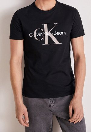 T-shirt en coton noir avec un grand logo « CK » blanc et gris, accompagné du texte « Calvin Klein Jeans » en dessous. Manches courtes et col rond.