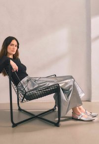 Czarny metalowy krzesło z kablowym wzorem. Modelka ma na sobie długą srebrną spódnicę i srebrne buty na obcasie z zapięciem.