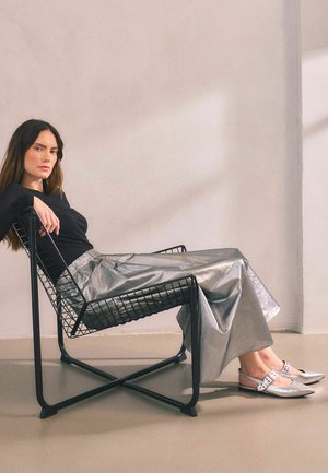 Chaise en fil métallique noir avec design géométrique. Le mannequin porte une longue jupe argentée et des chaussures à talons slingback argentées avec un détail de boucle.