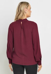 Blusa bordeaux con maniche lunghe, tessuto testurizzato, scollatura arricciata, dettaglio dei bottoni sui polsini e apertura a chiave sulla schiena.