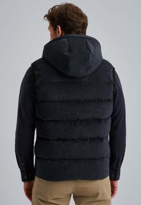 Mężczyzna w czarnej, pikowanej kamizelce z kapturem, noszący długosleeve'ową koszulę i beżowe spodnie, odwrócony tyłem na tle jasnego, jednolitego koloru.