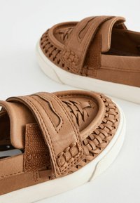 Bruna slip-on-skor med texturerade vävda detaljer, ett kardborreband och en vit gummisula. Släta material med rundad tådesign.