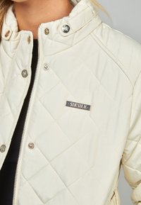 Quiltet creme jakke med høj krave, sølvfarvet hardware og et fremtrædende "SikSilk" logo prægede på en metalplade. Har trykknap- og lynlåslukning.