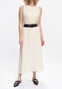 Vrouw in een mouwloze crèmekleurige midi-jurk met een brede zwarte riem en zwarte puntige flats met banden, staand op een witte achtergrond.