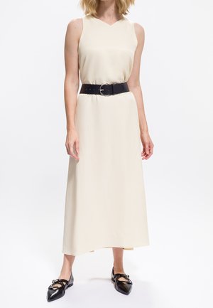 Femme portant une robe midi crème sans manches avec une large ceinture noire et des ballerines noires à talons plats, se tenant sur fond blanc.