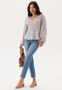 Blouse à manches longues florale en violet clair et crème avec des manches bouffantes ; portée avec un jean taille haute bleu et des sandales à talons beige.