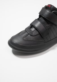 Sneaker nera con tomaia in pelle texturizzata, due cinturini in velcro e suola in gomma arrotondata. Presenta un logo discreto sulla linguetta.