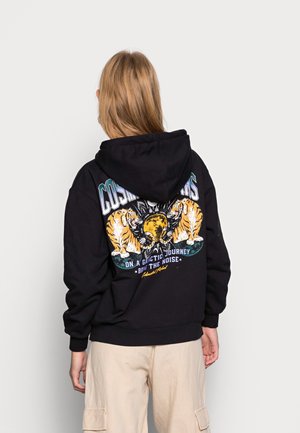 Person mit langen blonden Haaren, die einen schwarzen Hoodie mit zwei Tigern und einer gelben Planeten-Grafik trägt, beige Cargohosen, von hinten zur Kamera gewandt.