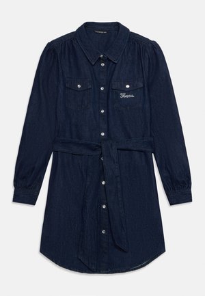 Długa koszulowa sukienka z ciemnoniebieskiego denimu z guzikami z przodu, kieszeniami na piersiach, materiałowym paskiem oraz logo "Guess" na lewej kieszeni wykonanym z małych kryształków.