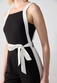 Černý overal s bílými bočními akcenty a nastavitelným pasem. Vyznačuje se širokými ramínky a čtvercovým výstřihem. Hladká textilie.