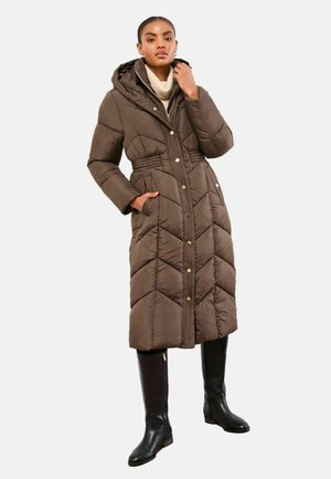 Cappotto imbottito lungo marrone con cappuccio, caratterizzato da un motivo a rombi, bottoni a pressione e tasche laterali. Indossato sopra stivali neri fino al ginocchio.