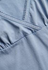 Close-up van lichtblauwe stof met decoratieve naden en geplooide stiksels, waarbij de textuur en details van de kledingconstructie worden benadrukt.