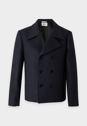 Homme portant un manteau croisé noir, une chemise rayée vert clair, un pantalon noir avec une ceinture marron et des baskets beiges, debout devant un fond uni.