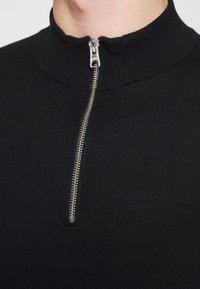 Primo piano di un top in maglia nera a coste con una cerniera metallica argentata zipparata a metà all'altezza dello scollo.