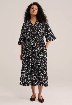 Frau mit lockigem Haar, die ein schwarzes Midikleid mit weißen abstrakten Blattmustern, halblangen Ärmeln und braunen flachen Schuhen trägt.