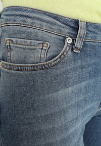 Bleka blå denimjeans med en frontfickdetalj, överstygn och metallknappstängning vid midjan, som visar en slät yta.