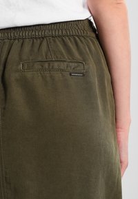 Pantalons vert olive avec une taille élastique, dotés d'une poche arrière en patch et d'un tissu lisse et texturé.