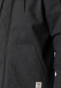 Mørkegrå hættetrøje med lynlås, frontlomme og lille hvid label med en elg-logo nær lommens nederste hjørne.