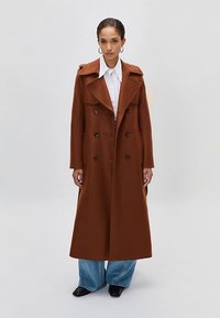 Žena v dlhom hnedom dvojradovom kabáte, s bielou košeľou, širokými modrými džínsami a čiernymi topánkami, postavená na jednolatom pozadí.