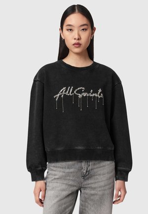 Jonge vrouw met stijl zwart haar draagt een zwarte trui met zilverversierde letters en lichtgrijze jeans op een effen achtergrond.