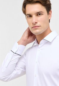 Hvid skjorte med sorte accenter på kraven og manchetterne. Har en slim fit og glat tekstur. Modellen justerer kraven på billedet.