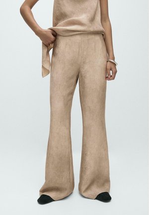Persona che indossa pantaloni beige a gamba larga e top senza maniche coordinato, con scarpe nere e braccialetto d'argento al polso destro.