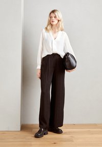 Blusa bianca a maniche lunghe con scollo a V, abbinata a pantaloni neri a vita alta e gamba larga. La modella tiene una borsa nera e indossa scarpe chunky.
