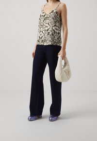 Top senza maniche con motivo floreale in oliva e crema, abbinato a pantaloni a gamba larga blu scuro. Tiene in mano una borsa color crema e indossa scarpe viola.