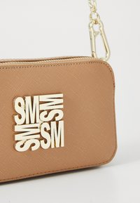 Pochette da polso marrone con texture, dotata di cerniera dorata e iniziali metalliche sovrapposte in oro "SM" sul fronte, attaccata a una catena dorata.