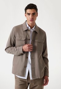 Jachetă din bumbac maro deschis, cu închidere cu nasturi, două buzunare la piept și un design cu guler, suprapusă peste o cămașă albă. Textură netedă.