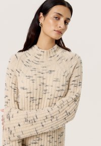 Beige Strickpullover mit hohem Kragen, geripptem Muster und schwarzen Sprenkeln, mit langen Ärmeln, die über die Handgelenke hinausreichen.