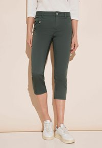 Pantalones recortados de color verde oscuro, confeccionados con una tela suave y elástica, que cuentan con un bolsillo con cremallera en la parte delantera y un cierre clásico de botón. Combinados con zapatillas blancas.