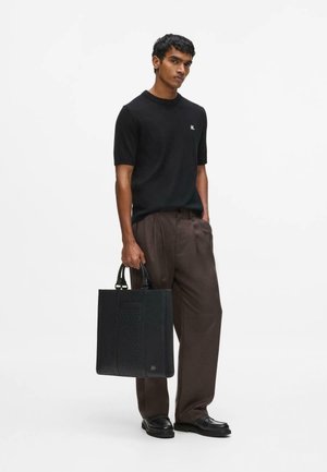 Man met zwart T-shirt met korte mouwen en bruine broek, houdend een zwarte draagtas, staand tegen een effen witte achtergrond.