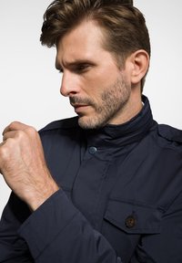 Granatowa kurtka z wysokim kołnierzem, teksturowana tkanina, zapięcie na guziki z przodu oraz kieszeń na piersi z guzikiem.