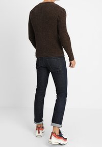 Personne portant un pull en maille marron, un jean foncé retroussé et des baskets blanches avec des détails noirs et rouges, debout devant un fond blanc.