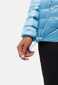 Lichtblauwe pufferjack met een gestructureerde stof, een getailleerde onderkant, verstelbare trekkoorden en een close-up van een hand die het koord vasthoudt.