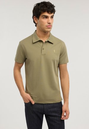 Junger Mann mit dunklen lockigen Haaren, trägt ein olivgrünes kurzärmeliges Poloshirt und dunkle Hosen, steht mit einer Hand in der Tasche.