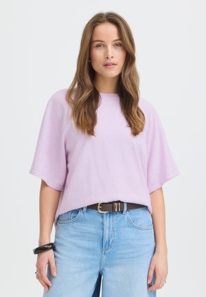 Młoda kobieta o długich brązowych włosach, ubrana w jasnofioletowy sweter z krótkim rękawem, jasnoniebieskie dżinsy, czarny pasek oraz czarną bransoletkę na lewym nadgarstku.