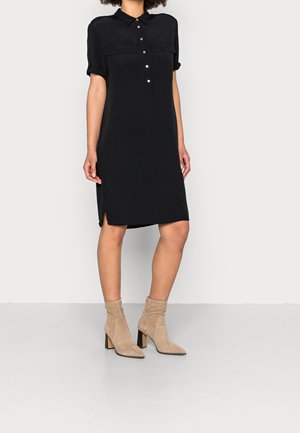 Mujer con vestido negro de manga corta abotonado con dos bolsillos en el pecho y botines beige con tacón, de pie contra un fondo liso.