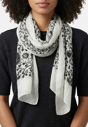 Mujer con una bufanda blanca con patrones florales y de paisley en negro, colocada sobre un suéter negro de manga corta.