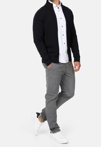 Cardigan lavorato a maglia nera da uomo con colletto alto, indossato su una camicia bianca con bottoni. Abbinato a pantaloni grigi e sneaker bianchi.