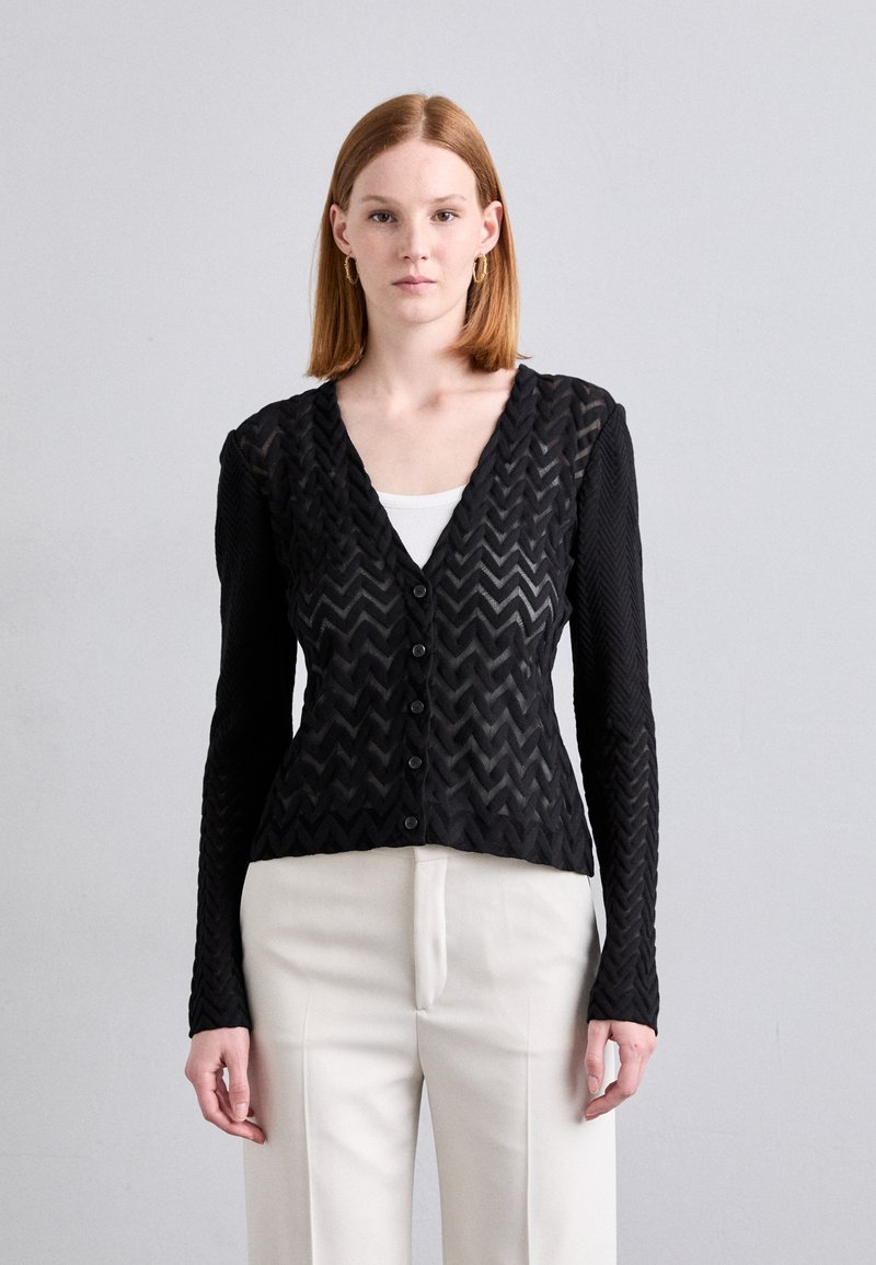 Cardigan noir texturé avec un col en V, un motif en chevrons, des manches longues et une fermeture à boutons sur le devant, superposé sur un haut blanc et associé à un pantalon beige.