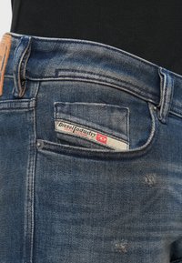 Τζιν παντελόνι με ξεθωριασμένο μπλε χρώμα, που διαθέτει δερμάτινο λογότυπο στην πλάτη, ετικέτα με την επιγραφή "Diesel Industry" και φθαρμένες λεπτομέρειες.